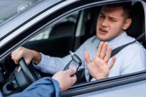 Inside the car, the driver reluctantly raises his hand while the officer outside presents a breathalyzer, mindful of California Breathalyzer Law and its implications.