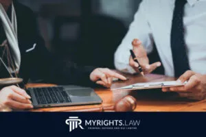 Two people in business attire sit at a desk with a laptop and documents, discussing paperwork. The logo 