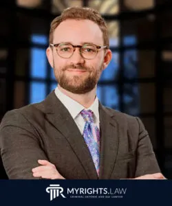 A man in a suit and tie stands with arms crossed, smiling, in front of a blurred background. The logo and text 