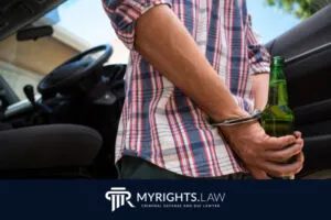 Una persona esposada que sostiene una botella de cerveza se encuentra junto a la puerta abierta de un automóvil, con 