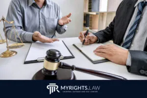 Dos personas están sentadas en un escritorio en Los Ángeles discutiendo asuntos legales con un abogado especializado en delitos menores; una escribe en un cuaderno mientras la otra gesticula. Un mazo y una balanza legal descansan sobre el escritorio.