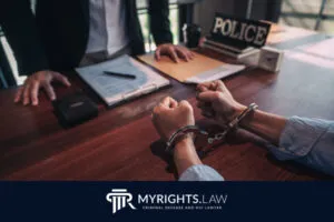 Una persona esposada se sienta en un escritorio frente a un oficial; papeleo policial y un cartel con la etiqueta