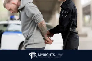 Un policía esposa a un hombre tatuado detenido junto a una patrulla. El logotipo y el texto... 