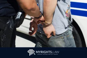 Un policía esposa a una persona con vaqueros y camiseta gris junto a una patrulla, lo que resalta la importancia de contar con un abogado penalista en Santa Ana. El logotipo y el sitio web del bufete aparecen en la parte inferior.