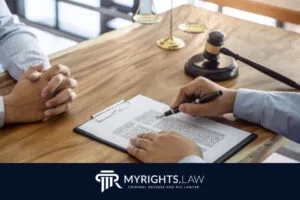 Dos personas se sientan en un escritorio de madera en Santa Ana discutiendo un documento legal con un abogado. Se ven un portapapeles, un bolígrafo, un mazo y una balanza. El logotipo de MyRights.Law aparece en la parte inferior, destacando su experiencia en defensa penal.