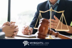 Two people sit at a desk, one taking notes while the other listens, with a scale of justice in view; the MyRights.Law logo is visible at the bottom, highlighting their Riverside criminal defense expertise.