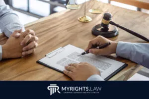 Dos personas se sientan en un escritorio de madera con un documento legal, un portapapeles, un bolígrafo y un mazo; un logotipo en la parte inferior dice “MYRIGHTS.LAW Abogado de Defensa Penal y DUI”, destacando la experiencia en las leyes de violencia doméstica de California.