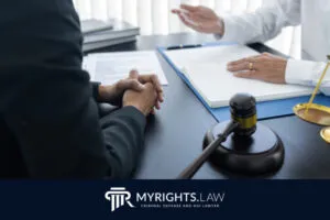 Dos personas se sientan en un escritorio con documentos legales, un mazo y una balanza de la justicia visible, discutiendo las leyes de violencia doméstica en California. Una gesticula mientras la otra escucha atentamente. El logo en la parte inferior dice: 