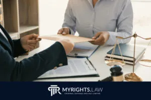 Dos personas vestidas de negocios intercambian un sobre a través de un escritorio que contiene documentos legales, un mazo y una balanza de justicia, lo que implica el impacto que un caso de DUI puede tener en el empleo; 