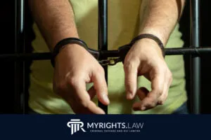 Una persona con una camisa amarilla se encuentra tras las rejas con las manos esposadas; el logotipo del abogado de defensa penal MYRIGHTS.LAW se muestra en la parte inferior.