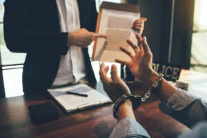 A person under arrest in handcuffs sits at a desk while another person in a suit points to a document, possibly discussing whether police did not read rights.