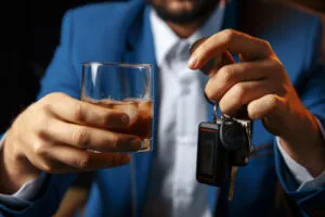 A person in a blue suit holds a glass of alcohol in one hand and car keys in the other, highlighting the danger of drinking and driving and illustrating the difference between DWI and DUI offenses.