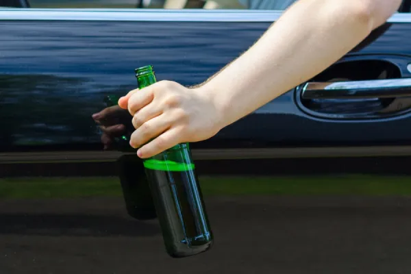 Una persona sostiene una botella de vidrio verde afuera de la ventana de un automóvil negro, arriesgándose a ser acusada de conducir bajo los efectos del alcohol (DUI) que podría afectar su empleo o sus perspectivas laborales.