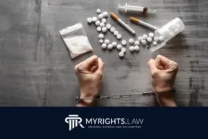 A pair of handcuffed hands with various drugs, syringes, pills, and powder on a table; 