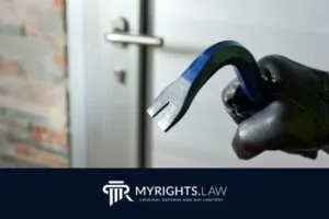 Una mano enguantada sostiene una palanca cerca de una puerta cerrada, lo que sugiere un allanamiento; el logotipo del abogado defensor penal My Rights Law Fullerton se muestra en la parte inferior.