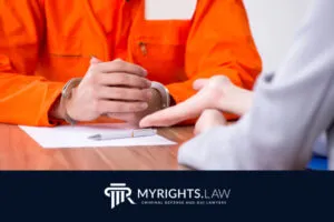 Person in orange prison uniform with handcuffs sits at a table, talking to an attorney in professional attire; MYRIGHTS.LAW logo at the bottom highlights expert criminal defense.