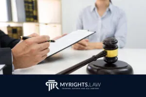 Un abogado que sostiene un portapapeles entrevista a otra persona en un escritorio con un mazo de juez en primer plano; se ven libros legales, el logotipo de MyRights.Law y materiales de defensa penal de Menifee.