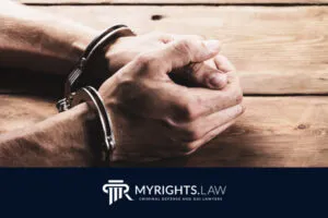 A person’s hands in handcuffs rest on a wooden table. Below, a logo reads 