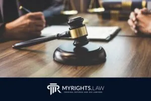 A gavel rests on a wooden table between two people in suits, with legal documents and books nearby; the