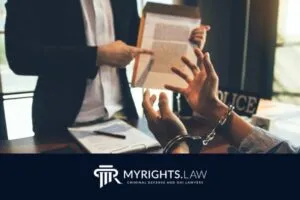 A person in handcuffs sits at a desk while another individual, possibly a Corona Criminal Defense Attorney, shows them a document; police items and paperwork are visible on the table.