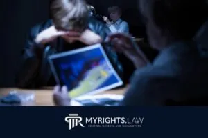 Dos personas se sientan en una mesa bajo una luz intensa mientras una le muestra una fotografía a la otra; una tercera persona se sienta al fondo. El logotipo de MyRights.Law y 