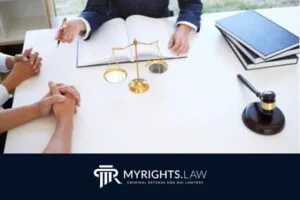 A criminal defense attorney meets with two clients at a desk with legal books, a scale of justice, and a gavel. The image includes the