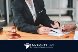 Two people sit at a desk discussing documents; one holds a pen, the other gestures with their hands. A gavel and legal scales are visible. Text reads 