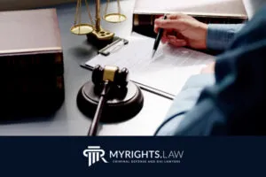 Persona que revisa y firma documentos legales en un escritorio con un mazo, una balanza de justicia y libros de leyes; el logotipo dice 