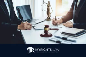 Two people in business attire sit at a desk with legal documents, a laptop, a gavel, and a Lady Justice statue—capturing the focus of a Murrieta criminal defense attorney. The logo 