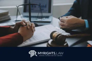 Dos personas se sientan frente a un escritorio con documentos legales, un mazo y una balanza de la justicia entre ellas, sugiriendo una consulta legal con un abogado penalista de Menifee. El logotipo de MYRIGHTS.LAW aparece en la parte inferior.