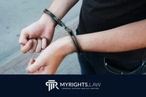 Una persona esposada se encuentra en una acera de Victorville con las manos al frente, sobre un logotipo de My Rights Law para servicios de abogados de defensa penal.