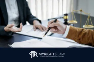 Dos personas están sentadas en un escritorio con documentos legales, un libro y una balanza de justicia; una sostiene un bolígrafo. 