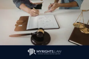 Two people discuss notes and charts at a desk with a gavel, scales of justice, and folders. Logo at bottom reads 