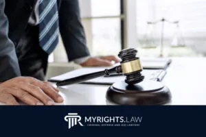 A person in a suit stands beside a judge’s gavel and scales of justice on a desk, with the 