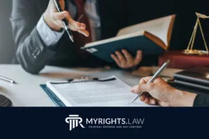 Two people in business attire review legal documents at a desk; one holds a pen while the other gestures with a book. Logo reads 