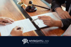 Dos personas sentadas en un escritorio en Rancho Cucamonga, revisan y firman documentos legales con un abogado cómplice; hay un mazo, una balanza y un portapapeles. El logotipo de MyRights.Law se muestra en la parte inferior.