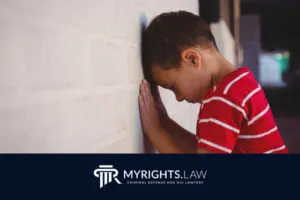 Young boy in a red striped shirt stands facing a white brick wall with his head resting against it. 