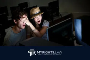 A woman in a hat covers a man's eyes as both look surprised at a computer screen in a dimly lit room with other computers. The 